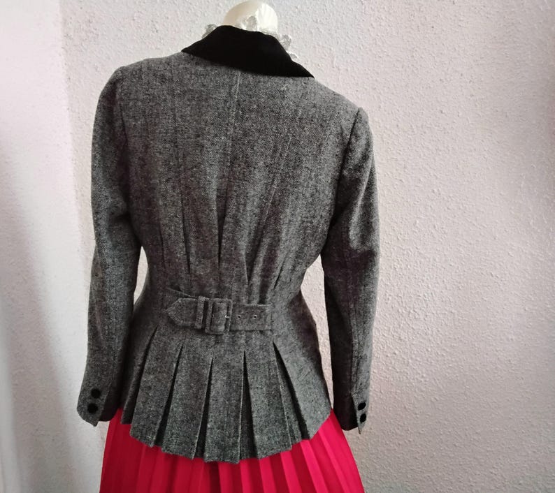 May include: A grey tweed jacket with a black velvet collar and a pleated peplum. The jacket has a belt at the waist and black buttons on the sleeves. The jacket is paired with a bright red skirt.