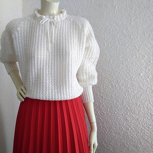 May include: White knitted sweater with a ruffled neckline and a red pleated skirt.