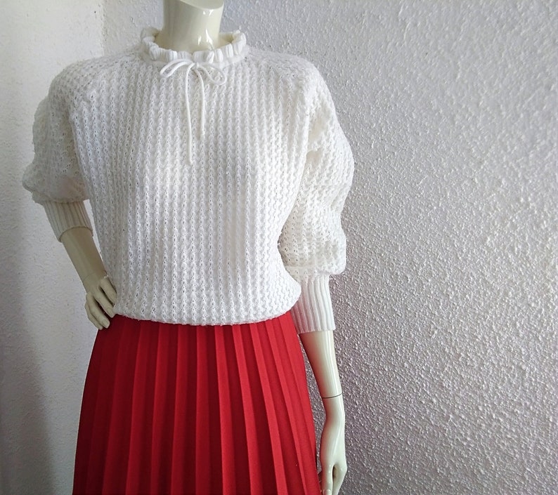 May include: White knitted sweater with a ruffled neckline and a red pleated skirt.