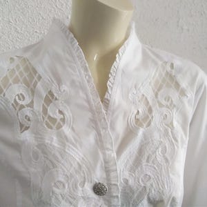 K&ouml;nnte beinhalten: Wei&szlig;es Hemd mit Kn&ouml;pfen, R&uuml;schenkragen und zarten Spitzendetails. Die Spitze ist ein durchsichtiger, wei&szlig;er Stoff mit Blumenmuster.