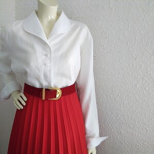 May include: A white long-sleeved button-up shirt with a wide collar and a red pleated skirt with a gold belt.