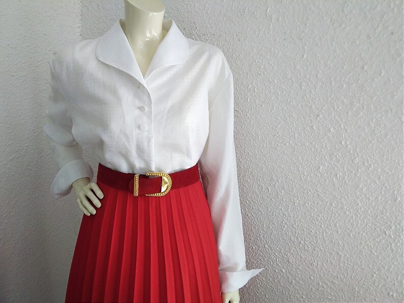 May include: A white long-sleeved button-up shirt with a wide collar and a red pleated skirt with a gold belt.