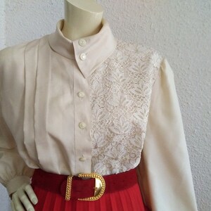 May include: A cream-colored blouse with a lace panel on the right side. The blouse has a high collar and long sleeves. A red belt with a gold buckle is worn over the blouse.