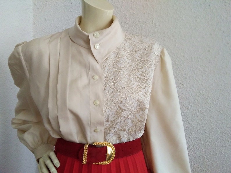 May include: A cream-colored blouse with a lace panel on the right side. The blouse has a high collar and long sleeves. A red belt with a gold buckle is worn over the blouse.