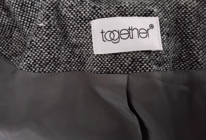 May include: Close-up of a garment's interior, showing a textured black and white tweed fabric. A grey lining is visible below the tweed. A white label with the word "together" is sewn into the tweed.