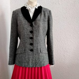May include: A grey tweed jacket with a black velvet collar and black buttons. The jacket is worn with a bright red pleated skirt. The mannequin is wearing a white blouse with a frilled collar.