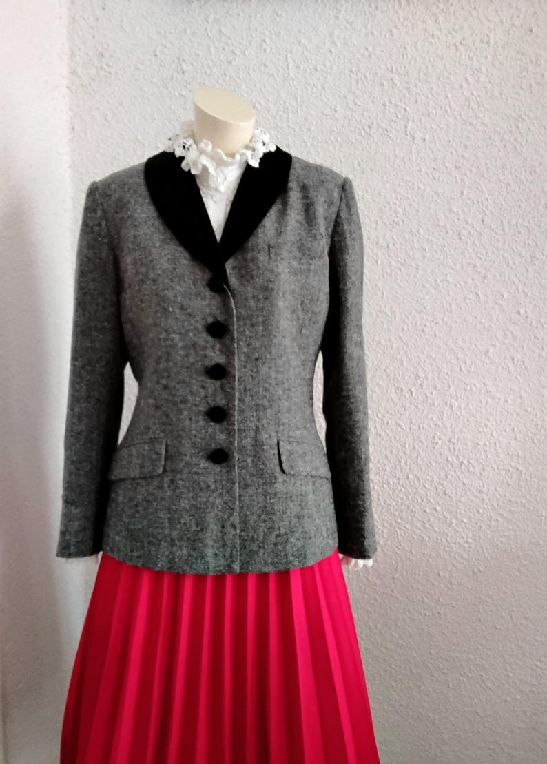 May include: A grey tweed jacket with a black velvet collar and black buttons. The jacket is worn with a bright red pleated skirt. The mannequin is wearing a white blouse with a frilled collar.