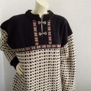 May include: A black and cream-coloured sweater with a geometric pattern. The yoke is black with decorative red and white embroidery and silver clasps. The sleeves and body feature a repeating square pattern.