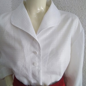 May include: A white button-down blouse with a wide collar and long sleeves. The blouse has a subtle textured pattern and is worn with a red belt with a gold buckle.
