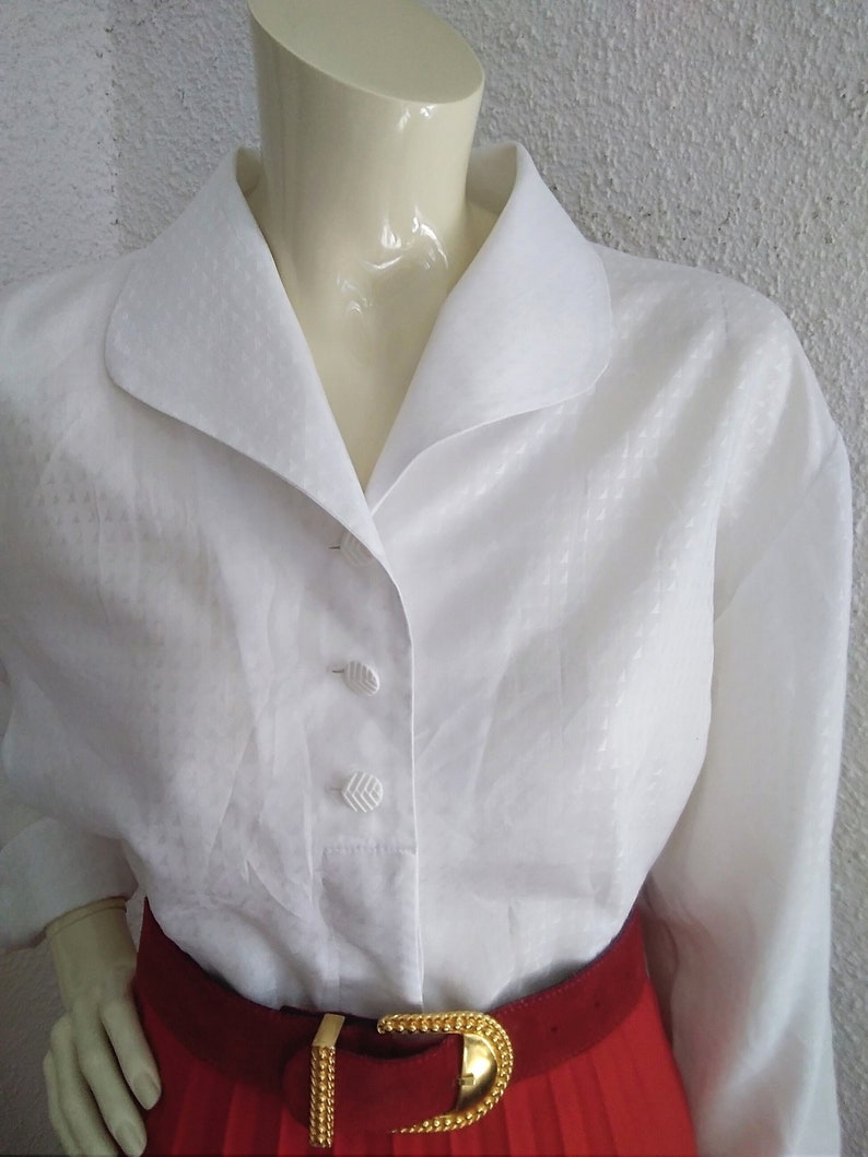May include: A white button-down blouse with a wide collar and long sleeves. The blouse has a subtle textured pattern and is worn with a red belt with a gold buckle.