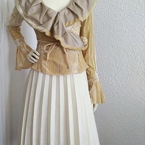 May include: A gold and beige blouse with a ruffled collar and long sleeves, paired with a white pleated skirt.