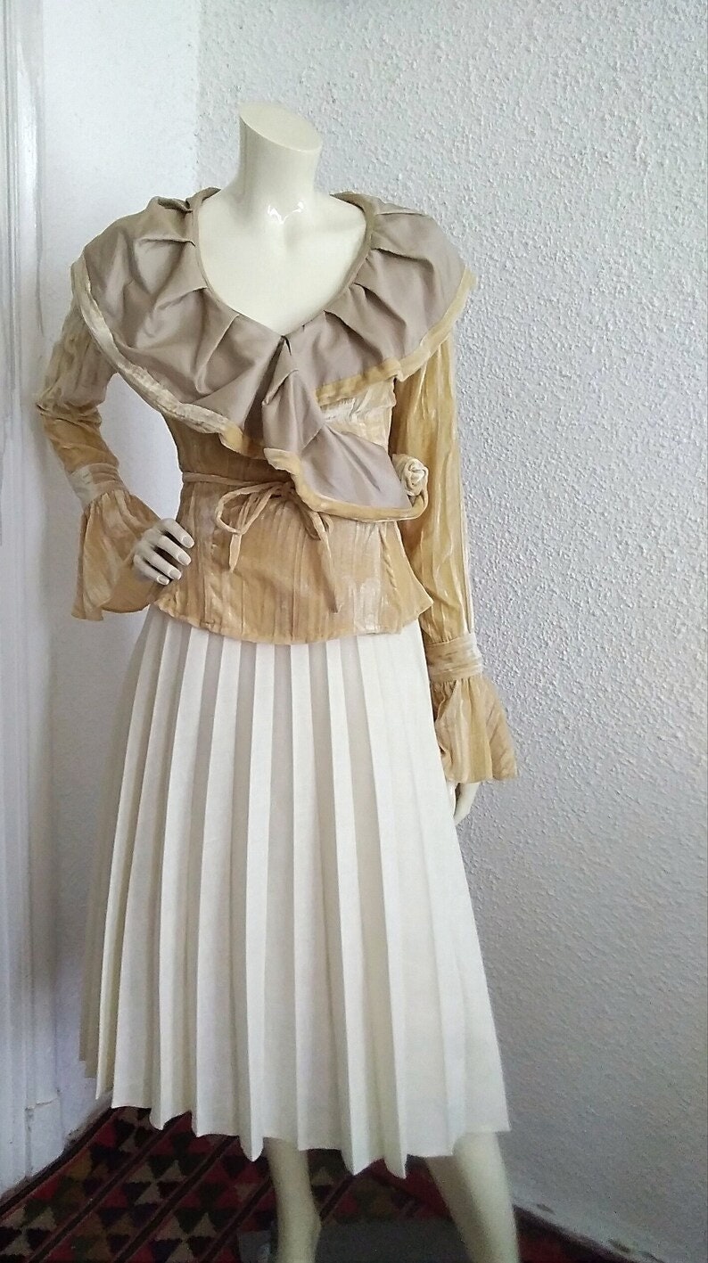 May include: A gold and beige blouse with a ruffled collar and long sleeves, paired with a white pleated skirt.