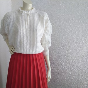 May include: A white crocheted sweater with a ruffled neckline and a red pleated skirt.
