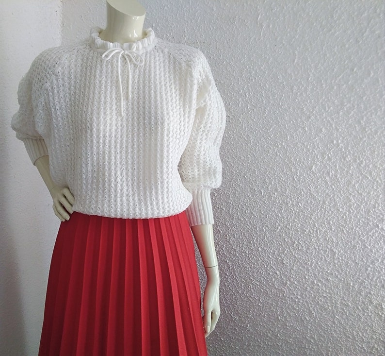 May include: A white crocheted sweater with a ruffled neckline and a red pleated skirt.