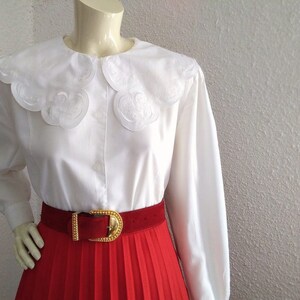 Puede incluir: Blusa blanca de manga larga con un gran cuello bordado con festones. La blusa se combina con una falda plisada roja y un cintur&oacute;n dorado.