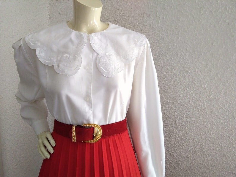 Puede incluir: Blusa blanca de manga larga con un gran cuello bordado con festones. La blusa se combina con una falda plisada roja y un cintur&oacute;n dorado.