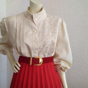 May include: A white blouse with a lace panel on the right side. The blouse has a high collar and long sleeves. The blouse is worn with a red pleated skirt and a gold belt.