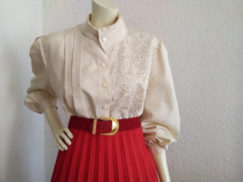 May include: A white blouse with a lace panel on the right side. The blouse has a high collar and long sleeves. The blouse is worn with a red pleated skirt and a gold belt.