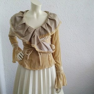 May include: A long-sleeved, beige velvet blouse with a large, ruffled collar and a tie at the waist. The blouse has bell sleeves and a V-neckline.