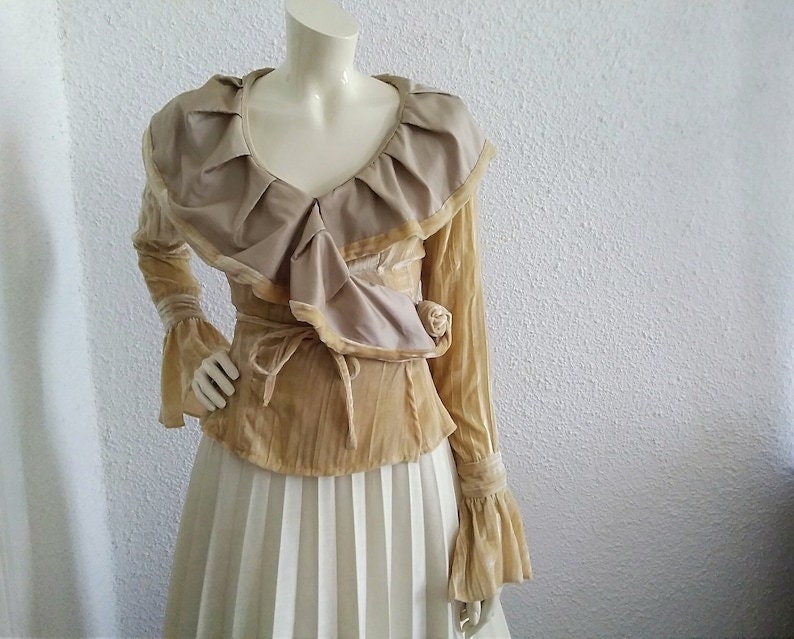 May include: A long-sleeved, beige velvet blouse with a large, ruffled collar and a tie at the waist. The blouse has bell sleeves and a V-neckline.