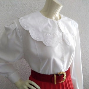 Puede incluir: Una blusa blanca de manga larga con un gran cuello blanco festoneado. El cuello tiene un delicado dise&ntilde;o bordado. La blusa se lleva con una falda plisada roja y un cintur&oacute;n dorado.