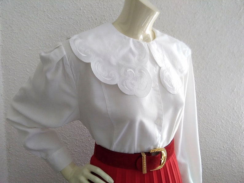Puede incluir: Una blusa blanca de manga larga con un gran cuello blanco festoneado. El cuello tiene un delicado dise&ntilde;o bordado. La blusa se lleva con una falda plisada roja y un cintur&oacute;n dorado.
