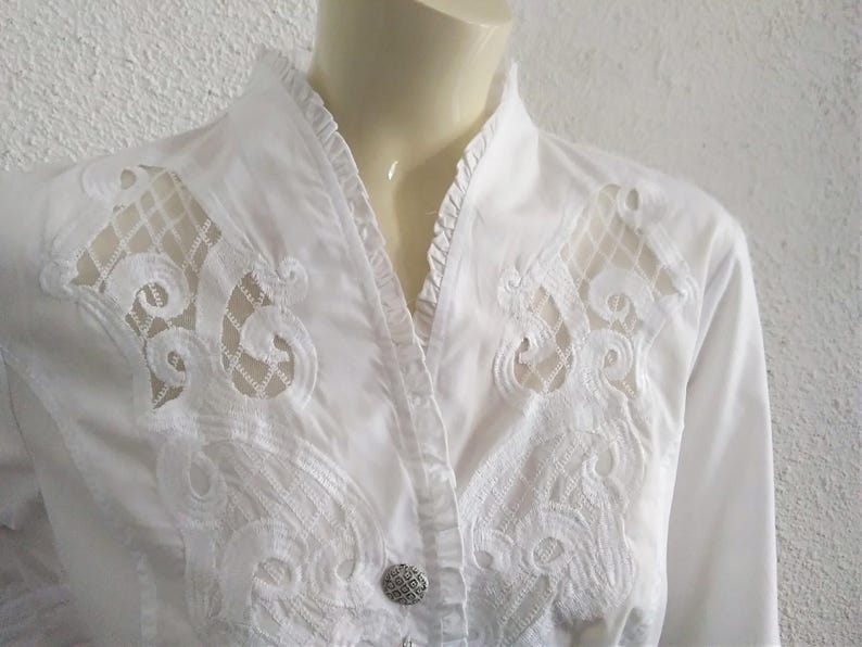 K&ouml;nnte beinhalten: Wei&szlig;es Hemd mit Kn&ouml;pfen und zarten Spitzendetails auf der Vorderseite. Die Spitze ist ein florales Muster mit einem durchsichtigen Design.