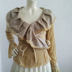 May include: A long-sleeved blouse with a large ruffled collar and a tie at the waist. The blouse is made of a soft, velvety fabric in a light beige colour.