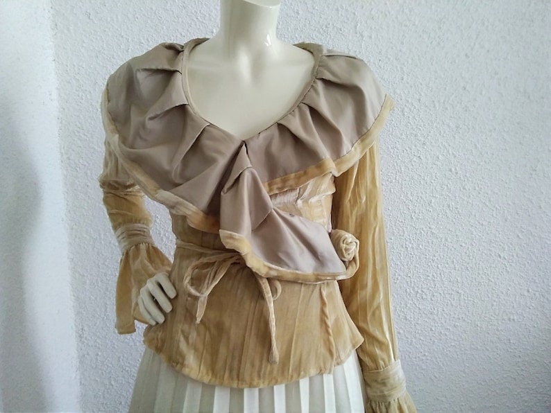 May include: A long-sleeved blouse with a large ruffled collar and a tie at the waist. The blouse is made of a soft, velvety fabric in a light beige colour.