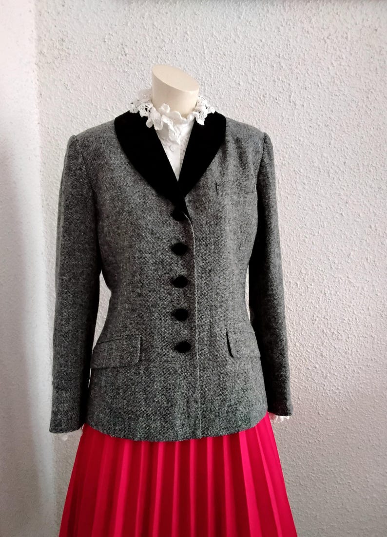 May include: A grey tweed jacket with a black velvet collar and black buttons. The jacket is worn with a white blouse with a lace collar and a bright pink pleated skirt. A classic, elegant outfit.