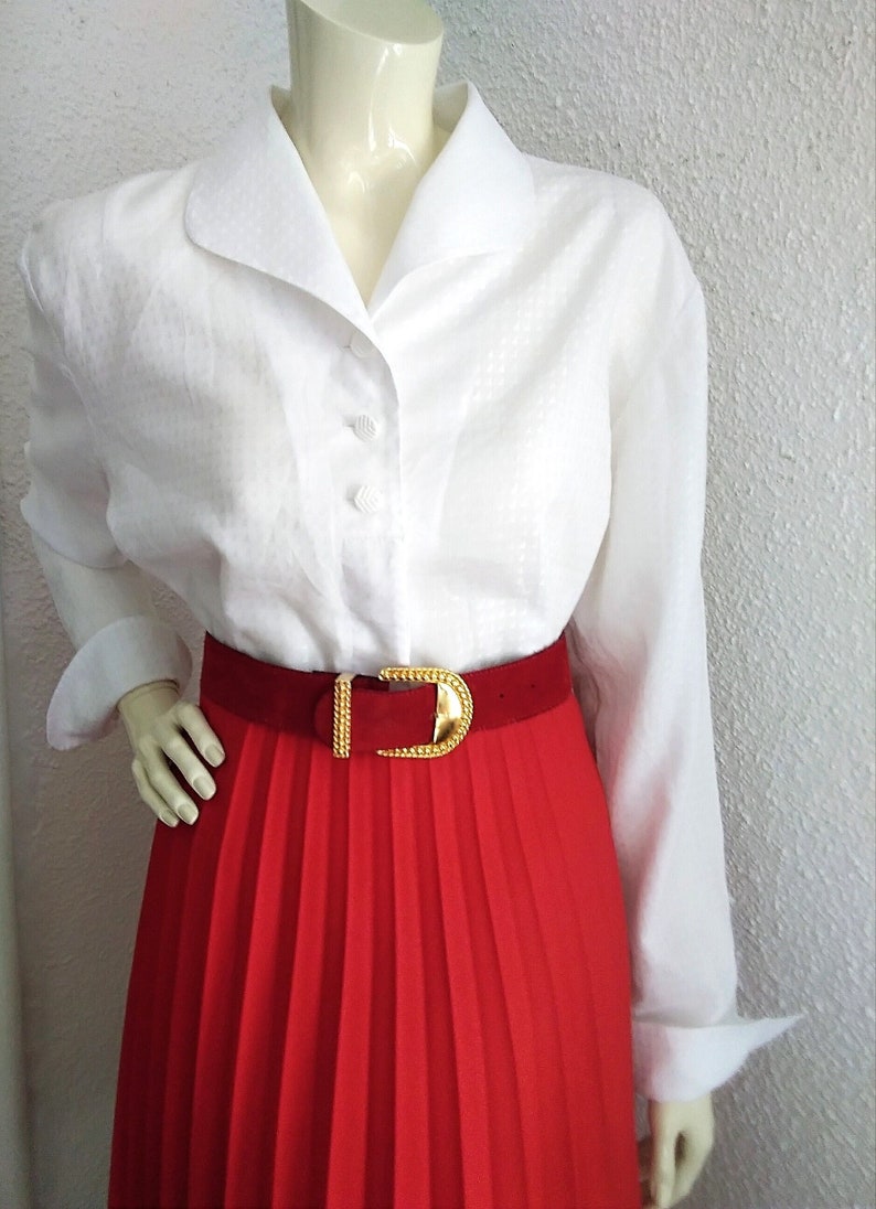 May include: A white button-down shirt with a wide collar and long sleeves. The shirt is worn with a red pleated skirt and a gold belt.