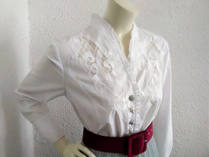 K&ouml;nnte beinhalten: Wei&szlig;e Bluse mit langen &Auml;rmeln, V-Ausschnitt und aufwendigen Spitzendetails auf der Vorderseite. Die Bluse hat einen burgunderfarbenen G&uuml;rtel.