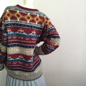 May include: A red, blue, and yellow patterned sweater with a crew neck. The sweater has a geometric design with stripes and a floral motif.