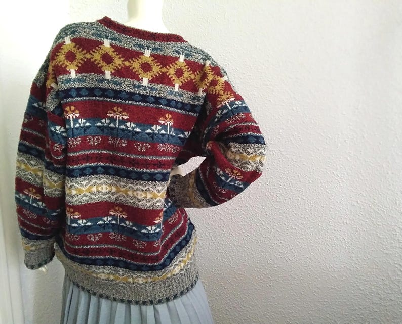 May include: A red, blue, and yellow patterned sweater with a crew neck. The sweater has a geometric design with stripes and a floral motif.