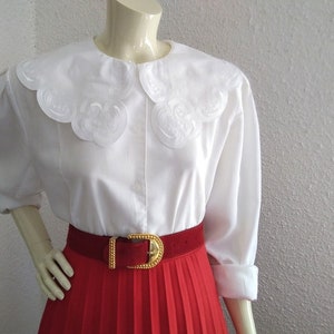 Puede incluir: Blusa blanca con un cuello grande y festoneado y mangas largas. La blusa se combina con una falda plisada roja y un cintur&oacute;n dorado.