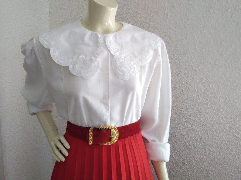 Puede incluir: Blusa blanca con un cuello grande y festoneado y mangas largas. La blusa se combina con una falda plisada roja y un cintur&oacute;n dorado.