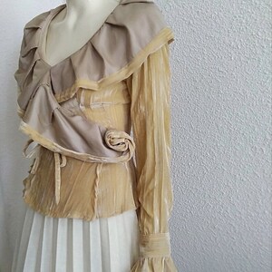 May include: A long-sleeved, beige velvet blouse with a ruffled neckline and a large, draped collar. The blouse has a fitted bodice and long, bell-shaped sleeves.