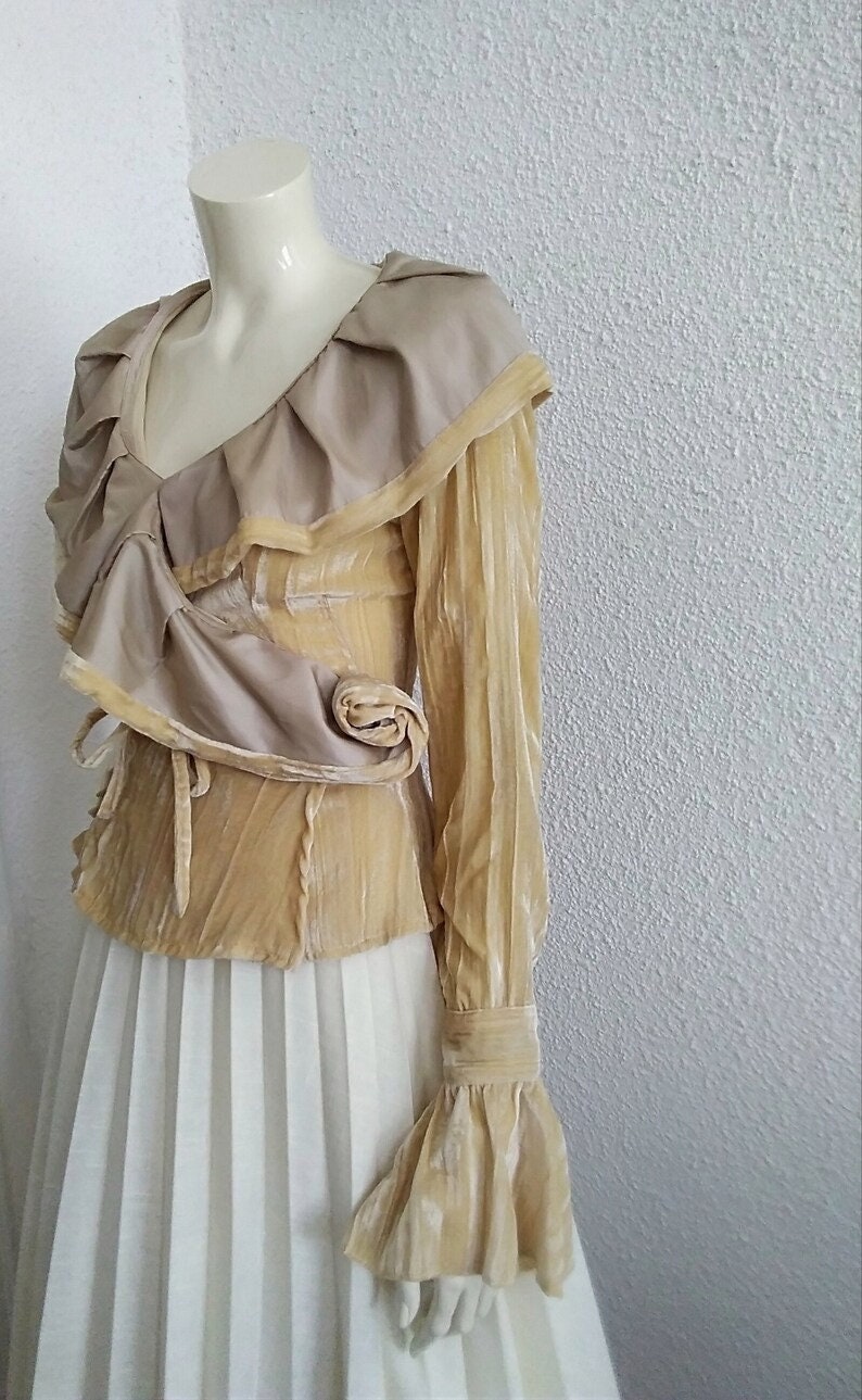 May include: A long-sleeved, beige velvet blouse with a ruffled neckline and a large, draped collar. The blouse has a fitted bodice and long, bell-shaped sleeves.