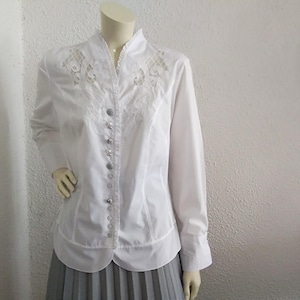 K&ouml;nnte beinhalten: Wei&szlig;e Bluse mit langen &Auml;rmeln, Stehkragen und zarten Spitzendetails auf der Vorderseite. Die Bluse hat einen Knopfleisten vorne und eine figurbetonte Silhouette.