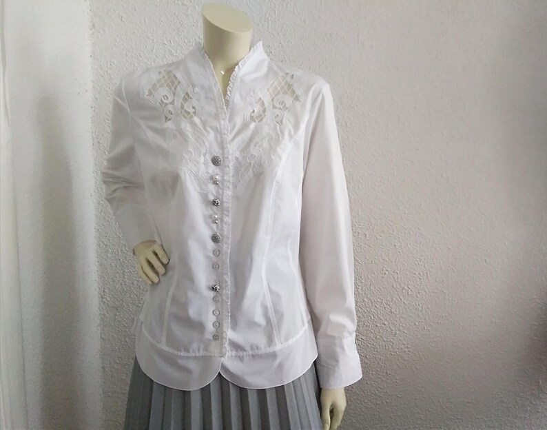 K&ouml;nnte beinhalten: Wei&szlig;e Bluse mit langen &Auml;rmeln, Stehkragen und zarten Spitzendetails auf der Vorderseite. Die Bluse hat einen Knopfleisten vorne und eine figurbetonte Silhouette.