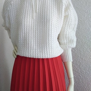 May include: A white knitted sweater with a ruffled collar and a tie at the neck. The sweater is paired with a red pleated skirt.