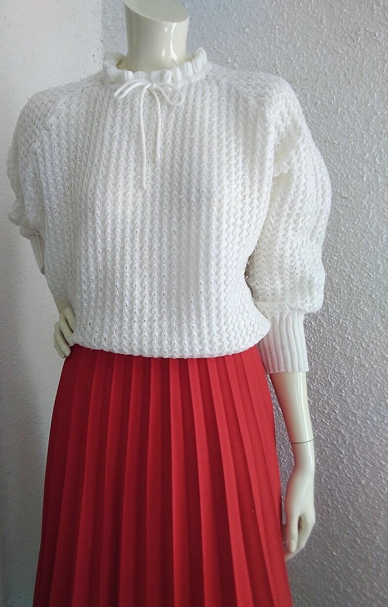 May include: A white knitted sweater with a ruffled collar and a tie at the neck. The sweater is paired with a red pleated skirt.