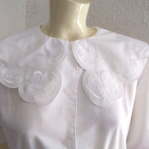 Puede incluir: Una blusa blanca con un cuello grande y festoneado. El cuello tiene un delicado dise&ntilde;o bordado.
