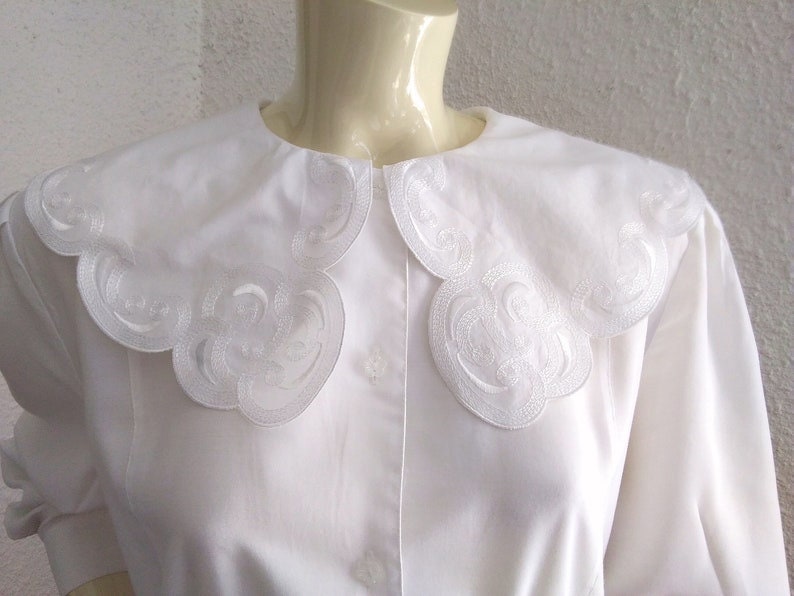 Puede incluir: Una blusa blanca con un cuello grande y festoneado. El cuello tiene un delicado dise&ntilde;o bordado.