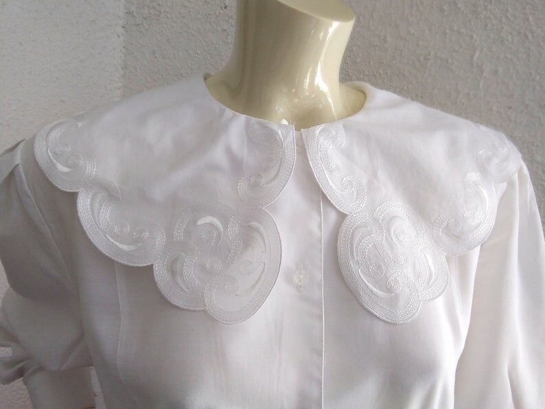 Puede incluir: Blusa blanca con un cuello grande festoneado con un dise&ntilde;o bordado. El cuello est&aacute; hecho de una tela blanca delicada y tiene un borde ligeramente fruncido.