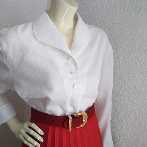 May include: White button-down blouse with a wide collar and a textured pattern. The blouse has long sleeves and is worn with a red pleated skirt and a gold belt.