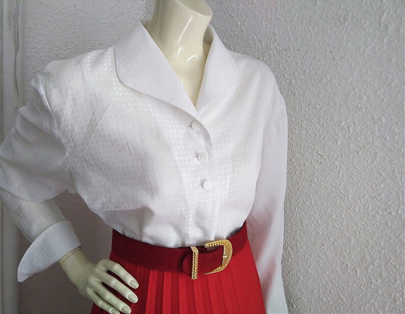 May include: White button-down blouse with a wide collar and a textured pattern. The blouse has long sleeves and is worn with a red pleated skirt and a gold belt.