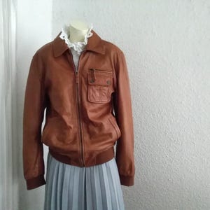 May include: A brown leather jacket with a zipper closure and a large pocket on the left side. The jacket has a ribbed collar and cuffs.