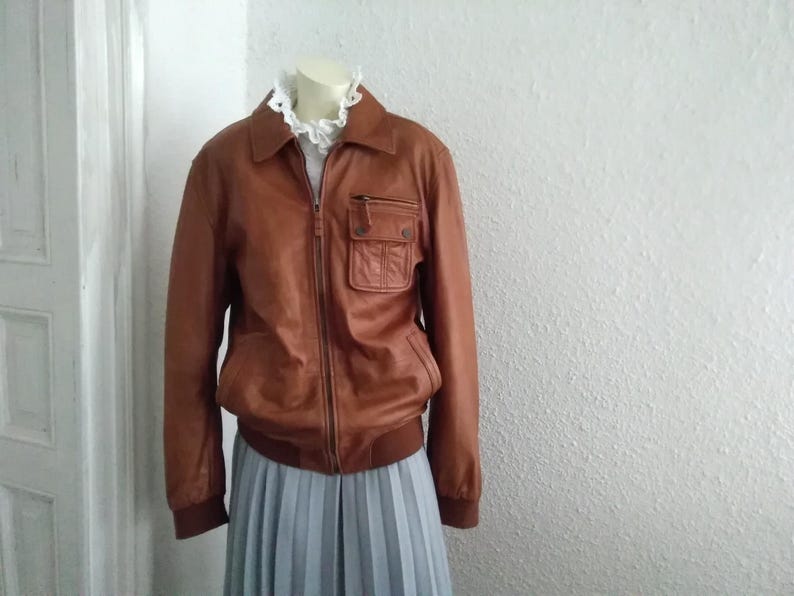 May include: A brown leather jacket with a zipper closure and a large pocket on the left side. The jacket has a ribbed collar and cuffs.