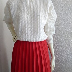 May include: A white knitted sweater with a ruffled neckline and a tie at the neck, paired with a red pleated skirt.